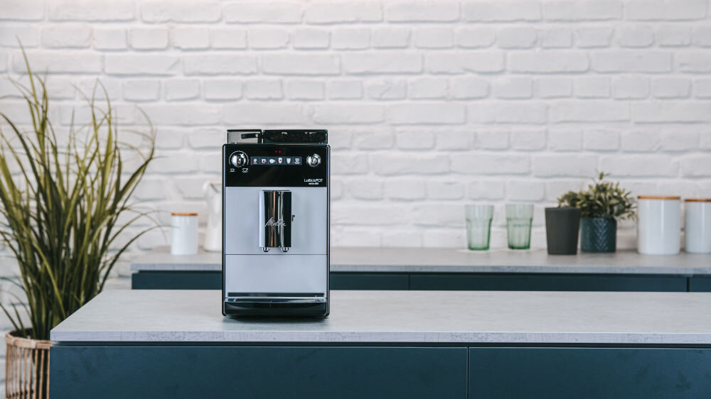 Na blacie kuchennym stoi srebrny ekspres do kawy z czarnymi elementami. W tle widoczna jest biała ceglana ściana oraz kilka roślin i szklanek.