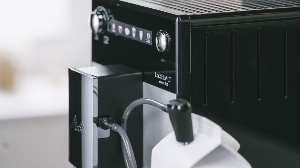 Zbliżenie na ekspres do kawy z widocznym panelem sterowania i napisem \'Latticia OT series 600\'. Obok znajduje się pojemnik z rurką do spieniania mleka.
