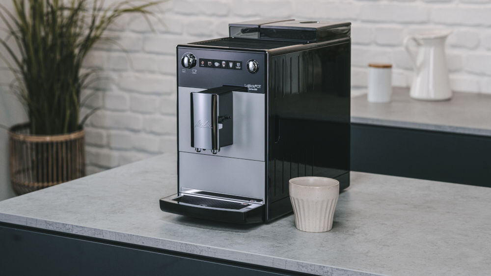 Na blacie kuchennym stoi srebrny ekspres do kawy z logo \'Melitta\' oraz filiżanka. W tle widoczna jest biała ceglana ściana i roślina w doniczce.