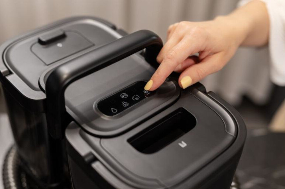 Osoba naciska przycisk na panelu sterowania odkurzacza piorącego. Panel zawiera kilka ikon wskazujących różne funkcje urządzenia.