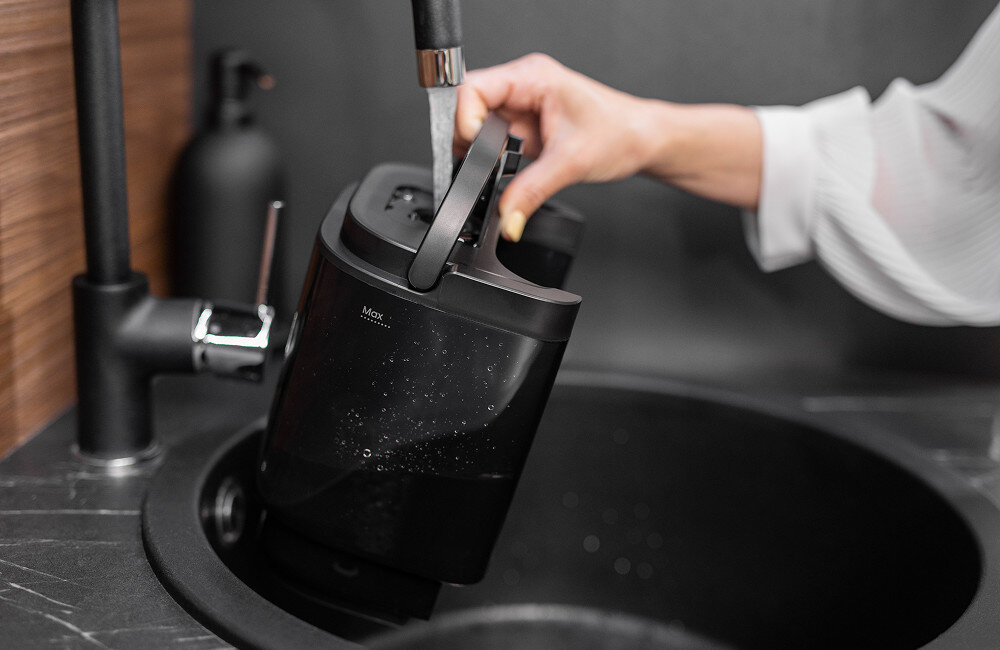 Osoba napełnia wodą czarny pojemnik w zlewie, na którym widoczny jest napis \'Max\'.
