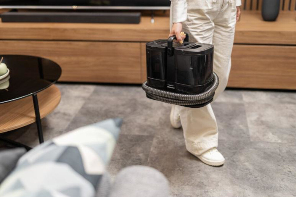 Osoba trzyma czarny odkurzacz piorący z wężem owiniętym wokół niego, stojąc na podłodze w pomieszczeniu z drewnianymi meblami. W tle widoczny jest stolik z filiżanką i talerzykiem.