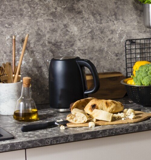Na blacie kuchennym znajduje się czarny czajnik elektryczny obok deski z pokrojonym chlebem i nożem. Obok stoi butelka z oliwą oraz miska z brokułem i papryką.