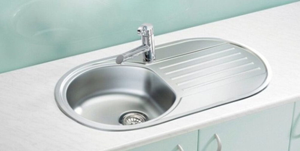 Na obrazie widoczny jest zlewozmywak dwukomorowy z jedną komorą głęboką i ociekaczem, wykonany ze stali nierdzewnej o satynowym wykończeniu. Zlewozmywak zamontowany jest na białym blacie, a nad nim znajduje się chromowana bateria.