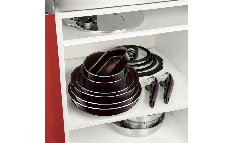 Na półce w szafce kuchennej znajduje się zestaw garnków i pokrywek w ciemnym kolorze oraz dwa uchwyty. W tle widoczna jest metalowa pokrywa.