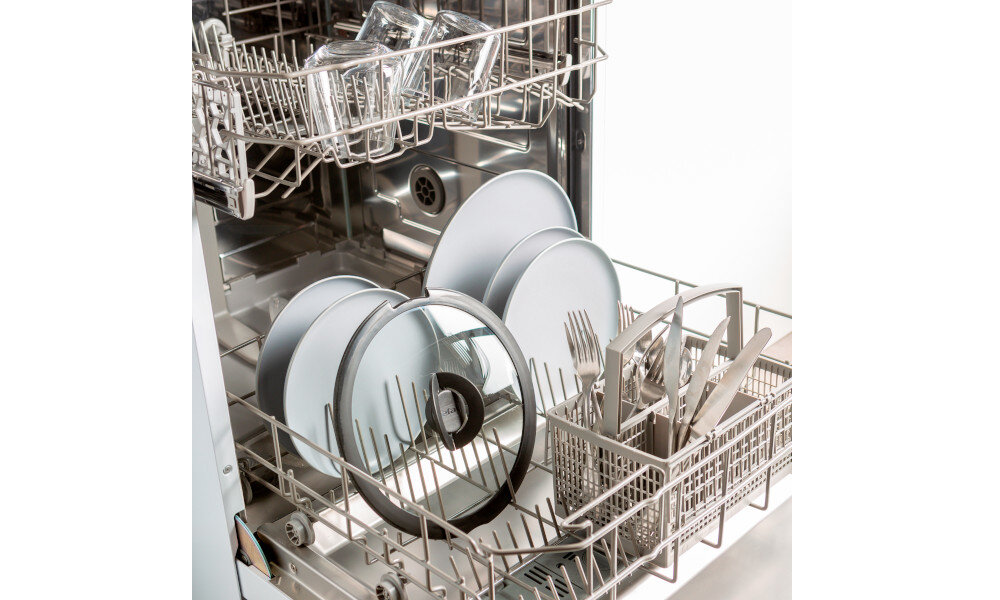 Wnętrze zmywarki z widocznymi pokrywami garnków, talerzami i sztućcami w koszyku. Pokrywy mają przezroczyste środki i metalowe obrzeża.
