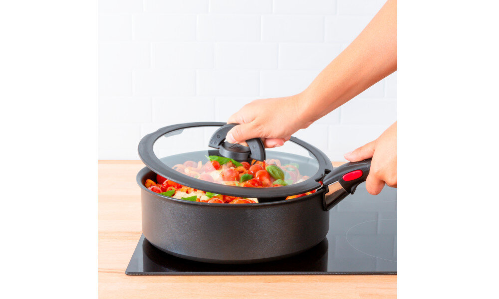 Osoba podnosi pokrywę z przezroczystym środkiem z garnka wypełnionego kolorowymi warzywami. Garnek stoi na płycie indukcyjnej w kuchni z jasnymi kafelkami w tle.