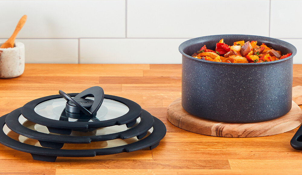 Na drewnianym blacie kuchennym znajduje się garnek z potrawą oraz zestaw trzech czarnych pokryw z przezroczystym środkiem. W tle widoczna jest biała ściana z płytek oraz drewniana łyżka w pojemniku.