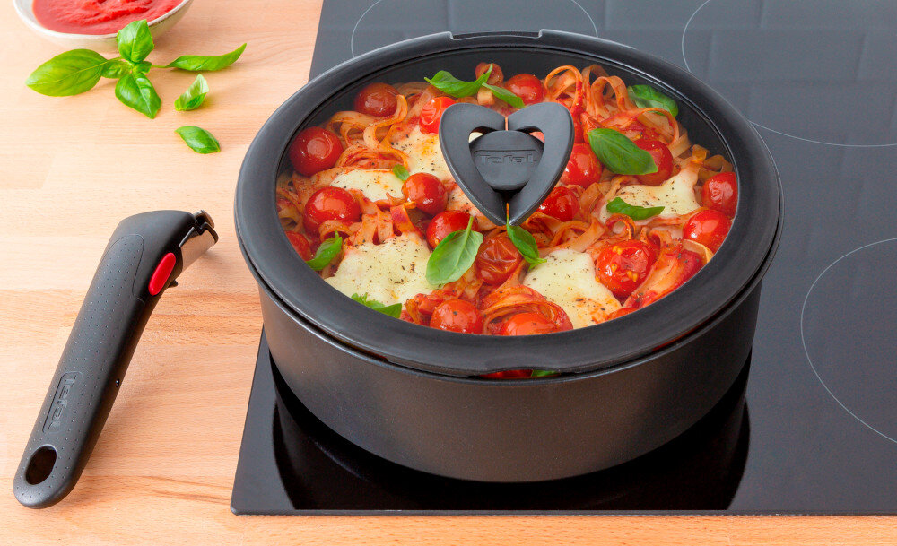 Na kuchence indukcyjnej stoi czarny garnek z przezroczystą pokrywą, wypełniony potrawą z pomidorami i ziołami. Obok leży odłączana rączka z czerwonym przyciskiem, a na pokrywie widoczne jest logo TEFAL.