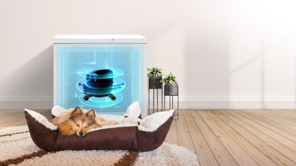 Na drewnianej podłodze stoi zamrażarka z widocznym niebieskim hologramem wewnątrz. Przed nią leży pies w brązowo-białym legowisku, a obok znajdują się dwie rośliny w doniczkach na stojaku.