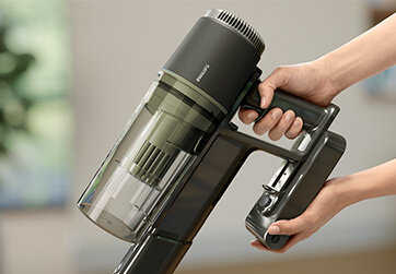 Osoba trzyma odkurzacz ręczny z widocznym logo \'PHILIPS\' na obudowie. Odkurzacz ma przezroczysty pojemnik na kurz i ciemną obudowę.