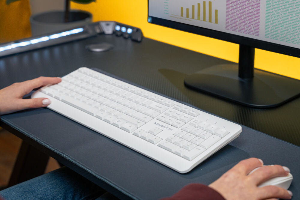Na biurku znajduje się biała klawiatura z napisem \'xquantum\' oraz myszka. W tle widoczny jest monitor z wykresami i tekstem na żółtym tle.