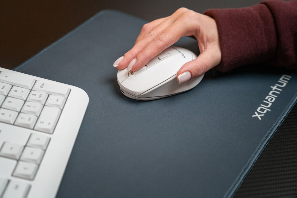 Na obrazie widoczna jest dłoń trzymająca białą mysz komputerową na ciemnej podkładce z napisem \'xquantum\'. Obok znajduje się fragment białej klawiatury.