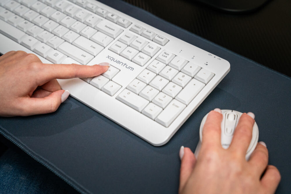 Na obrazie widoczna jest biała klawiatura z napisem \'xquantum\' oraz myszka, które są używane przez osobę. Klawiatura i myszka znajdują się na ciemnej podkładce.