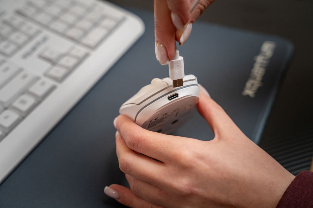 Osoba trzyma białą mysz komputerową i podłącza do niej kabel USB. Na podkładce pod mysz widoczne jest logo \'media expert\'.