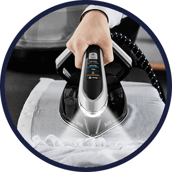 Osoba prasuje białą tkaninę przy użyciu żelazka z generatorem pary marki Tefal. Na żelazku widoczne są przyciski i wskaźniki, a z jego spodu wydobywa się para.