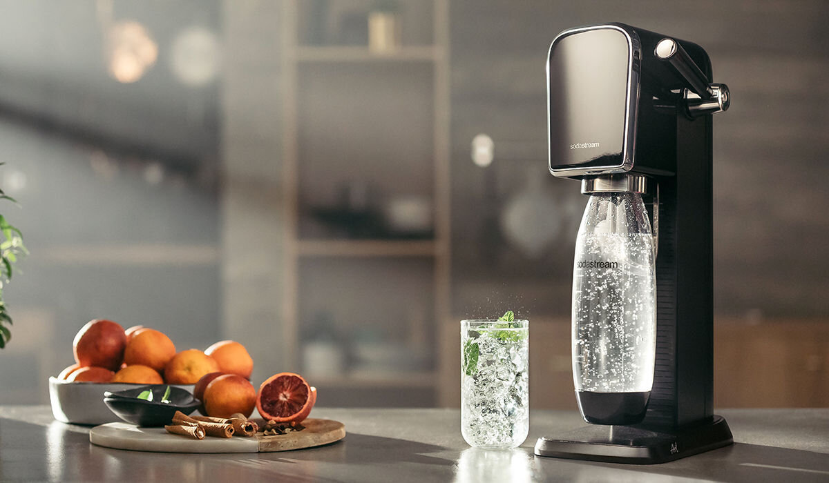 Na blacie kuchennym stoi czarny saturator z przezroczystą butelką wypełnioną gazowaną wodą. Obok znajduje się szklanka z napojem oraz miska z owocami i przyprawami na desce do krojenia.