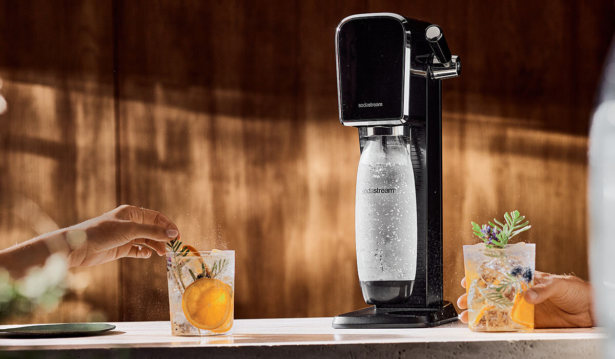 Czarny saturator SODASTREAM Art stoi na blacie, obok dwóch szklanek z napojami i dekoracjami. Na urządzeniu widoczne jest logo \'sodastream\'.