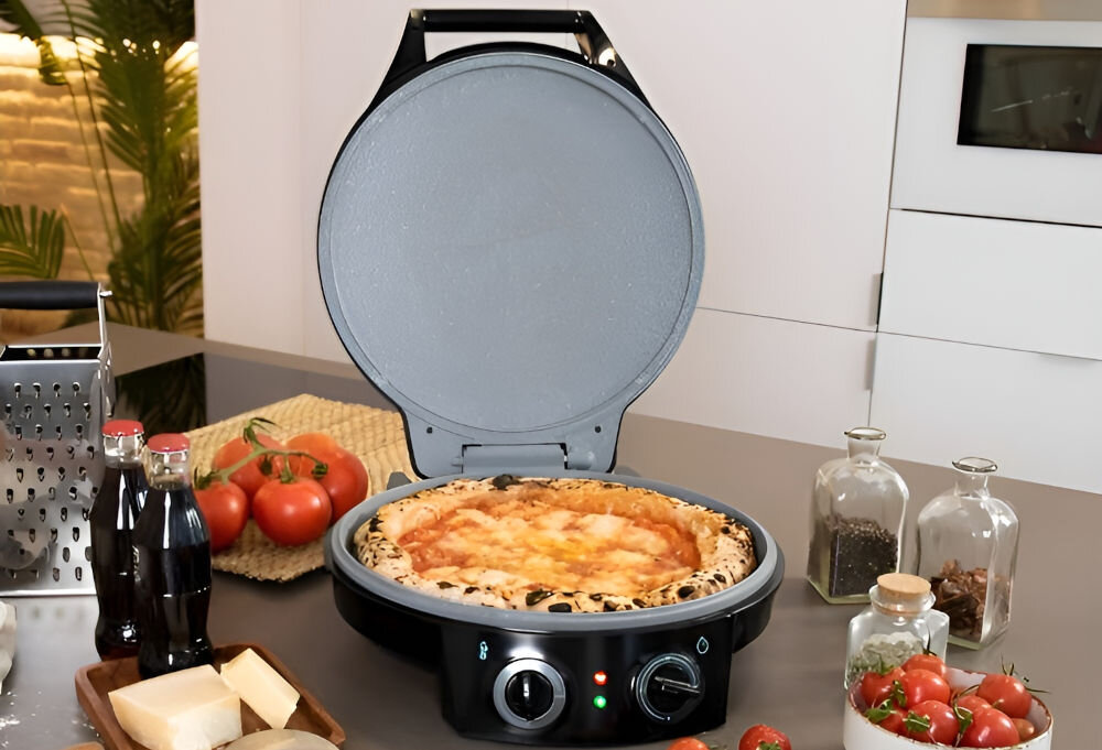 Na blacie kuchennym znajduje się otwarty piec do pizzy z gotową pizzą w środku. Obok leżą pomidory, ser, przyprawy w słoikach oraz butelki z ciemnym płynem.