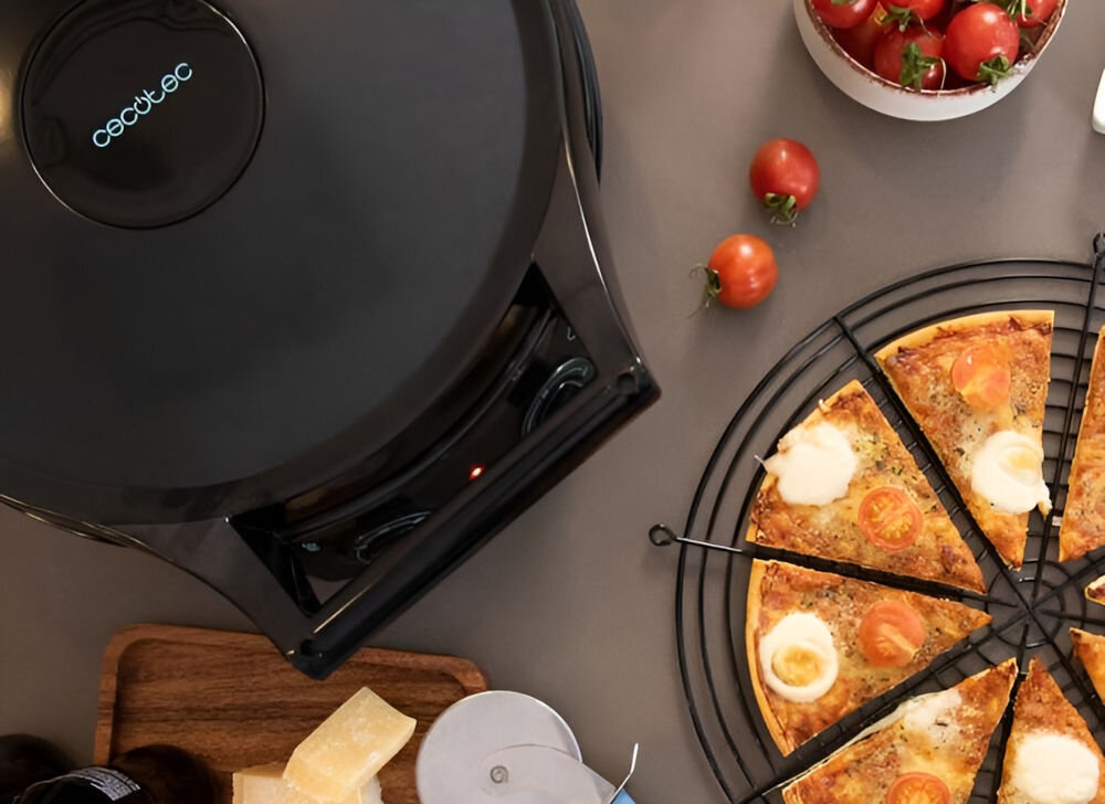 Na obrazie widoczny jest czarny piec do pizzy z logo \'cecotec\' oraz pokrojona pizza na metalowej kratce. Obok znajdują się pomidory i kawałki sera na drewnianej desce.