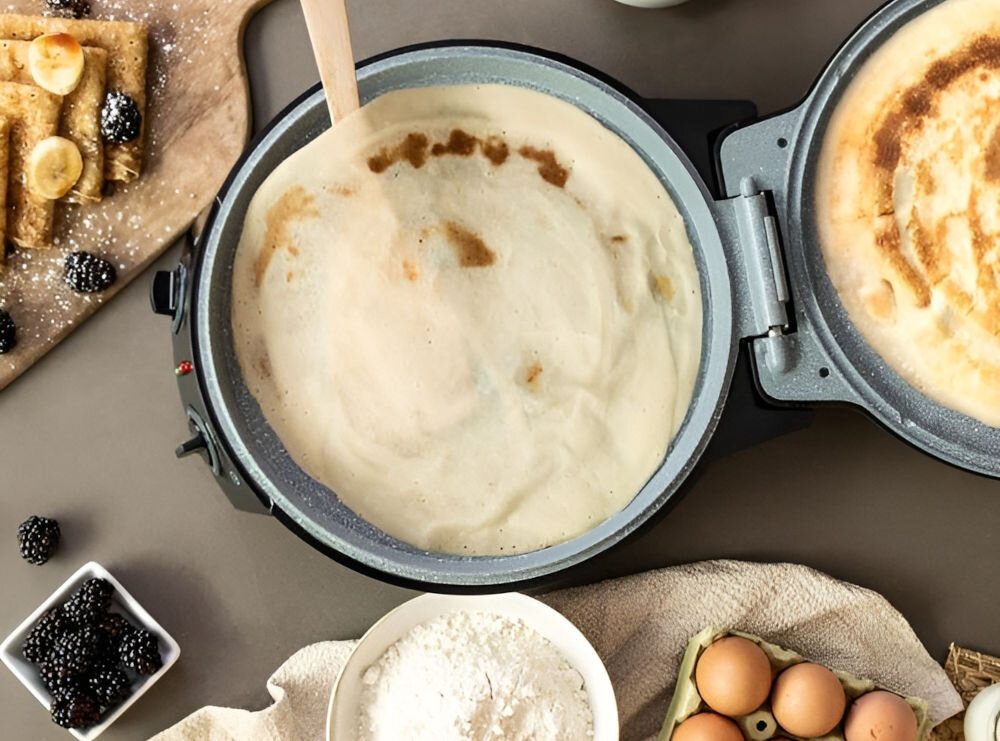 Na obrazie widoczny jest otwarty piec do pizzy z ciastem w środku. Wokół znajdują się składniki takie jak jajka, mąka, jeżyny i naleśniki z bananami.