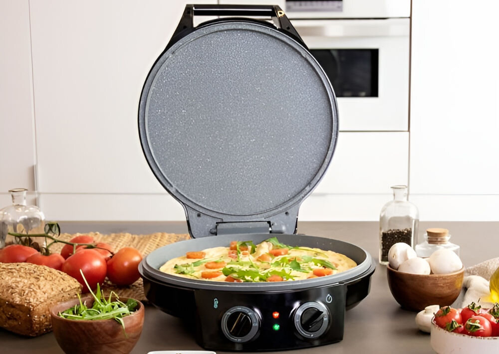 Na blacie kuchennym znajduje się otwarty piec do pizzy z widoczną pizzą w środku. Wokół pieca leżą różne składniki, takie jak pomidory, pieczarki i zioła.