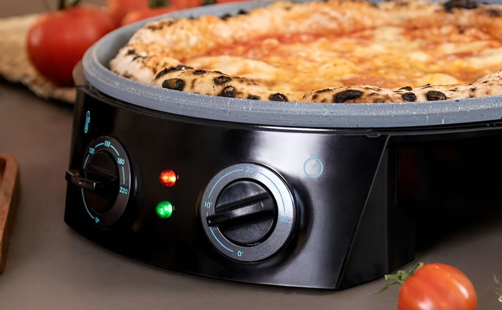 Na zdjęciu widać czarny piec do pizzy z dwoma pokrętłami i dwoma kontrolkami świetlnymi. Na piecu znajduje się pizza z widocznymi składnikami, a w tle leżą pomidory.