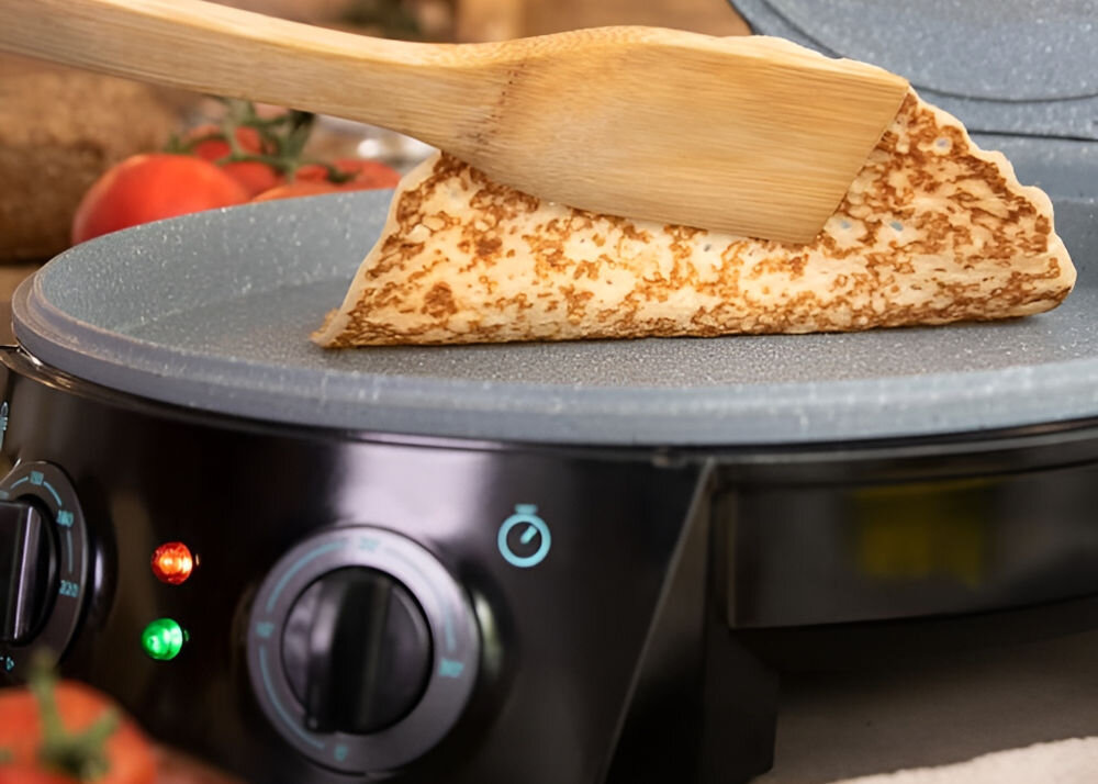 Na zdjęciu widać czarny piec do pizzy z szarą powierzchnią do pieczenia, na której znajduje się naleśnik podnoszony drewnianą łopatką. Na panelu pieca widoczne są dwa pokrętła z oznaczeniami temperatury i czasu oraz świecące kontrolki.