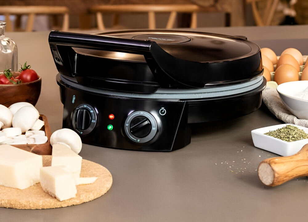 Czarny piec do pizzy z dwoma pokrętłami i trzema kontrolkami na panelu przednim stoi na blacie kuchennym. Wokół znajdują się składniki do pizzy, takie jak ser, pieczarki, pomidory i jajka.