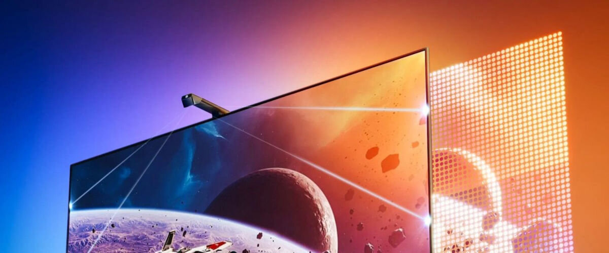 Telewizor z wyświetlanym obrazem kosmosu, z planetami i asteroidami, otoczony kolorowym podświetleniem LED. Tło jest gradientowe, przechodzące od niebieskiego do pomarańczowego.