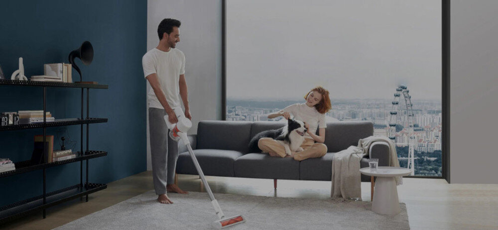 Osoba używa odkurzacza na dywanie w nowoczesnym salonie, podczas gdy inna osoba siedzi na kanapie z psem. W tle widoczne jest duże okno z widokiem na miasto.