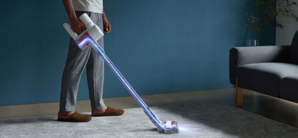 Osoba używa bezprzewodowego odkurzacza na dywanie w jasnym pokoju. Odkurzacz emituje niebieskie światło wzdłuż rury ssącej.