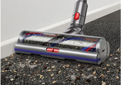 Na obrazie widoczna jest głowica odkurzacza z przezroczystą osłoną, która zbiera zanieczyszczenia z ciemnej powierzchni. Głowica ma czerwone elementy i jest przymocowana do metalowej rury.