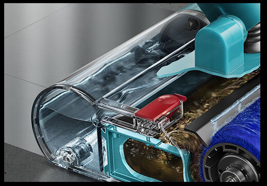 Zbliżenie na przezroczystą część odkurzacza z widocznym mechanizmem i niebieską szczotką. W tle widać fragment podłogi z szarych płytek.