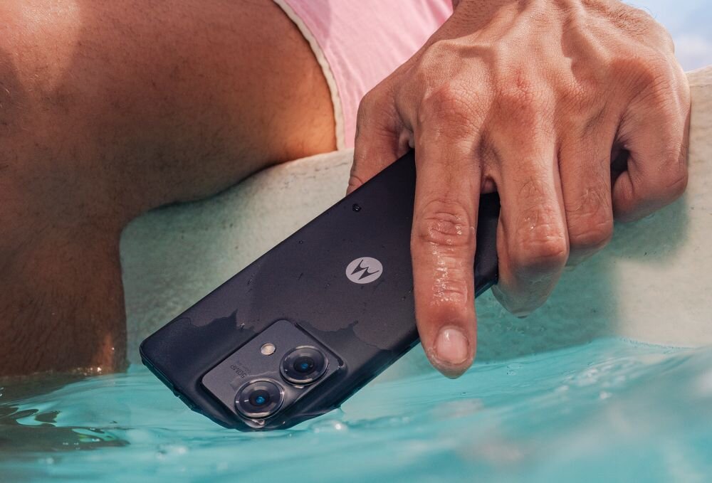 Osoba trzyma czarny smartfon z logo \'M\' nad wodą. Telefon ma dwa obiektywy aparatu na tylnej obudowie.