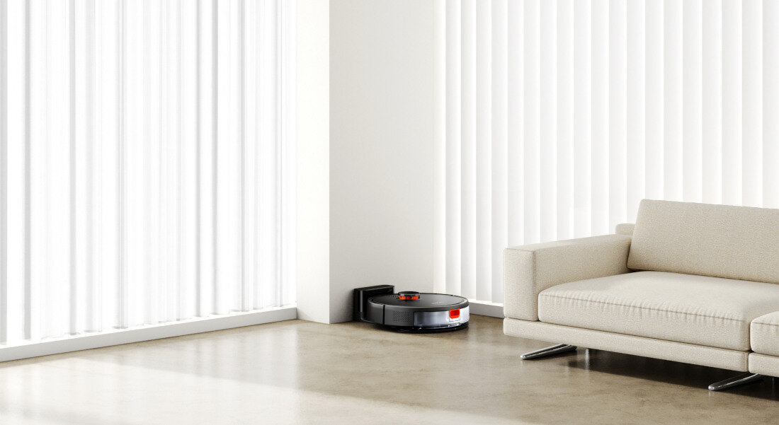 Na podłodze w jasnym pomieszczeniu znajduje się czarny robot sprzątający przy stacji dokującej. Obok stoi jasna sofa, a w tle widać pionowe żaluzje na oknie.