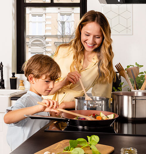 Kobieta i dziecko gotują na płycie ceramicznej w jasnej kuchni. Na blacie widoczne są garnki, patelnia z jedzeniem oraz deska z warzywami.