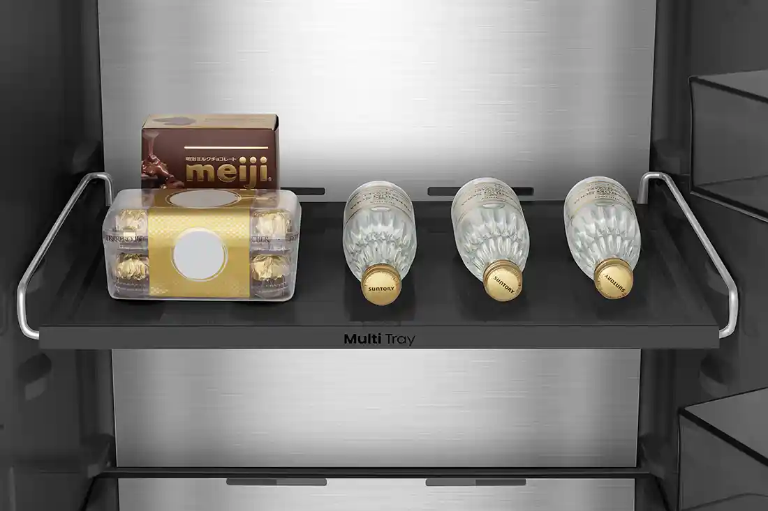 Wnętrze lodówki z półką oznaczoną jako \'Multi Tray\', na której znajdują się pudełko czekoladek \'meiji\', opakowanie pralinek oraz trzy butelki wody \'Suntory\'.