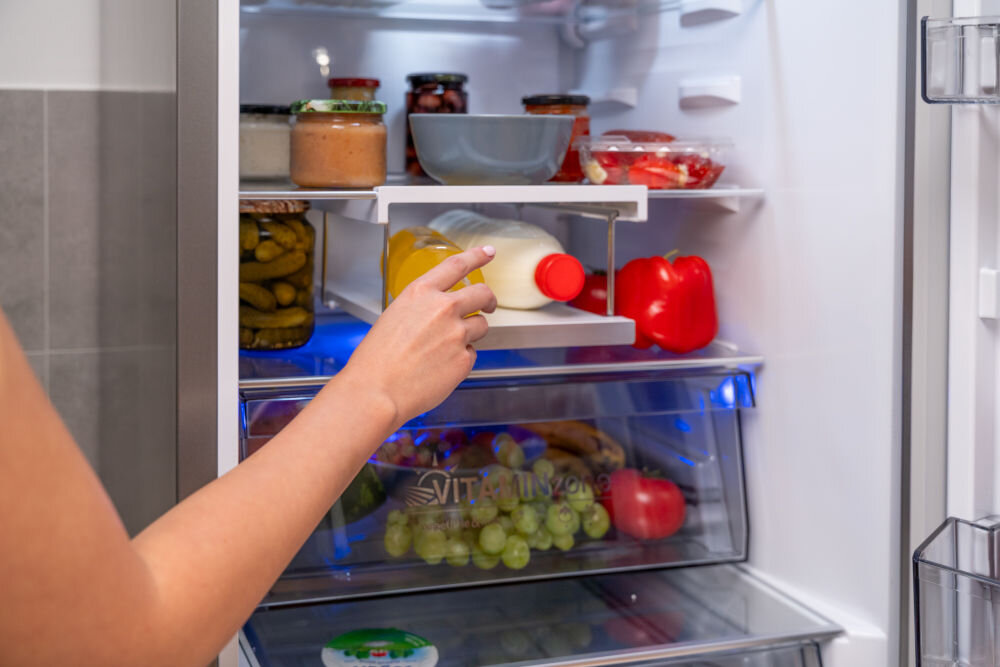 Otwarta lodówka z różnorodnymi produktami spożywczymi, w tym słoikami, butelkami i warzywami. Osoba sięga po butelkę z półki wewnątrz lodówki.