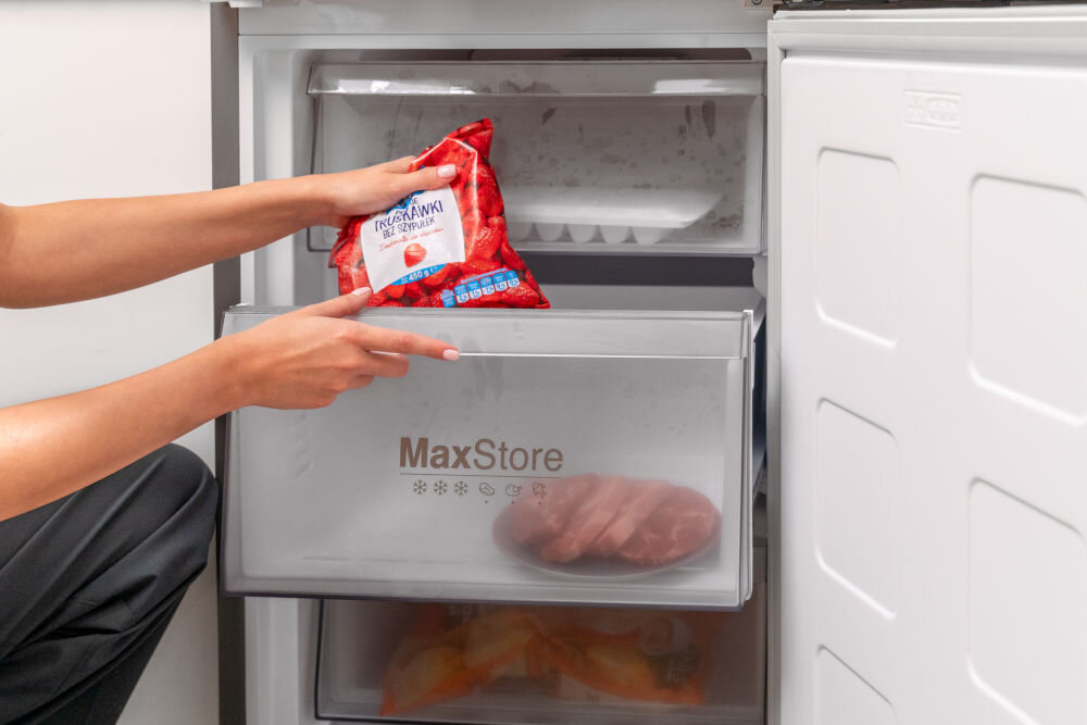 Osoba wkłada opakowanie mrożonych truskawek do zamrażarki z szufladą oznaczoną napisem \'MaxStore\'. Wewnątrz szuflady widoczne są zamrożone produkty.