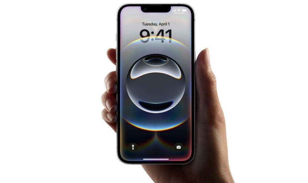 Osoba trzyma smartfon z wyświetlaczem pokazującym godzinę 9:41 i datę Tuesday, April 1. Na ekranie widoczna jest abstrakcyjna grafika z okręgami.