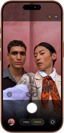 Ekran smartfona wyświetla aplikację aparatu z widocznymi przyciskami do robienia zdjęć i nagrywania wideo. W tle widać różową ścianę.