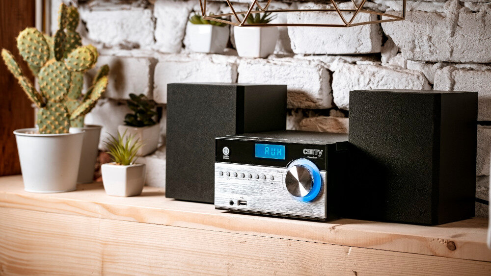 Wieża audio z wyświetlaczem pokazującym \'AUX\', otoczona dwoma głośnikami, stoi na drewnianej półce. W tle widoczne są białe cegły i kilka doniczek z roślinami, w tym kaktus.