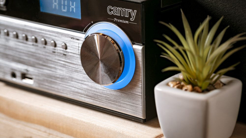 Na zdjęciu widoczna jest część wieży stereo z dużym pokrętłem otoczonym niebieskim światłem. Obok urządzenia stoi mała roślina w białej doniczce, a na wyświetlaczu widoczny jest napis \'AUX\'.