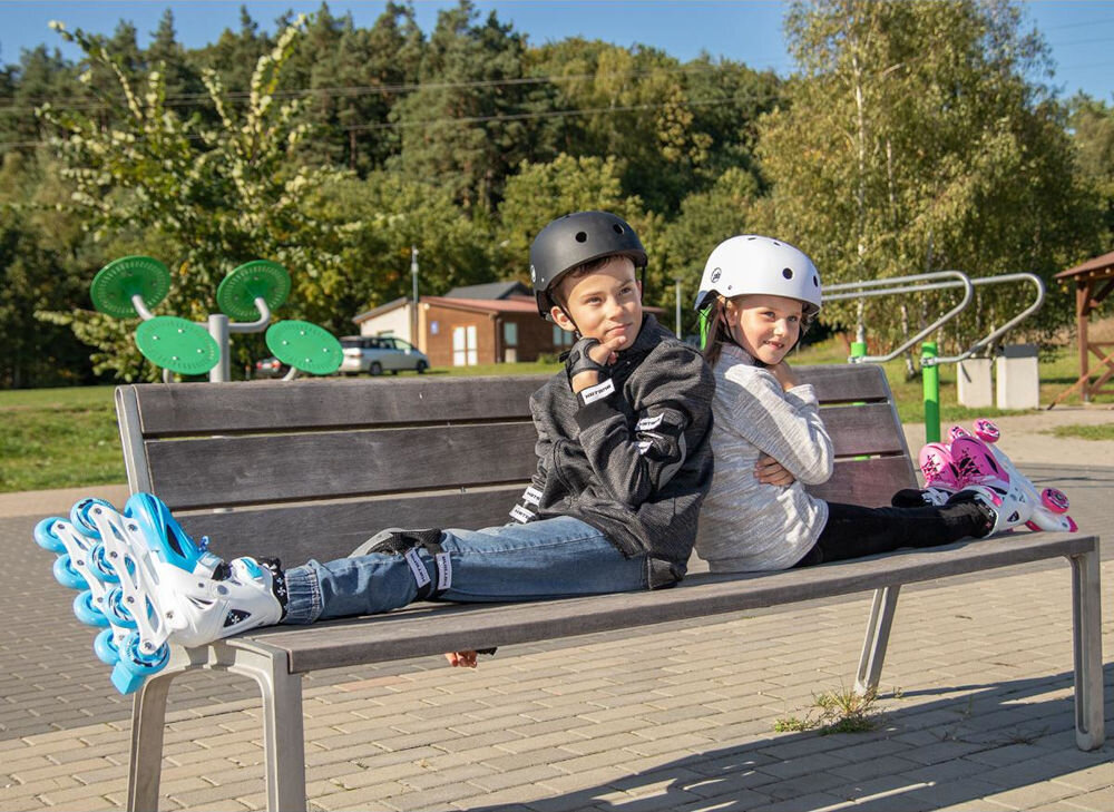 Dwoje dzieci siedzi na ławce w parku, mają na sobie kaski i łyżworolki. Łyżworolki są w kolorach niebieskim i różowym.