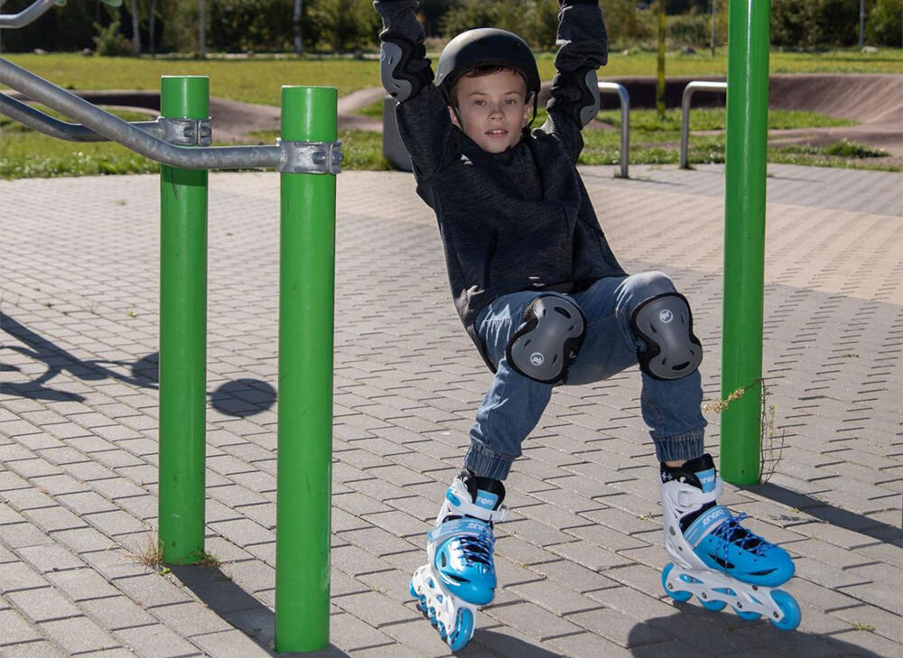 Dziecko w kasku i ochraniaczach na kolana i łokcie wisi na drążku na placu zabaw, mając na nogach niebiesko-białe łyżworolki. W tle widać zielone trawniki i betonową nawierzchnię.