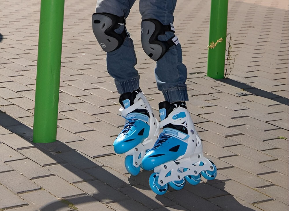 Osoba nosi niebiesko-białe łyżworolki z czterema kółkami, stojąc na chodniku. Na kolanach widoczne są ochraniacze, a w tle znajdują się zielone słupki.