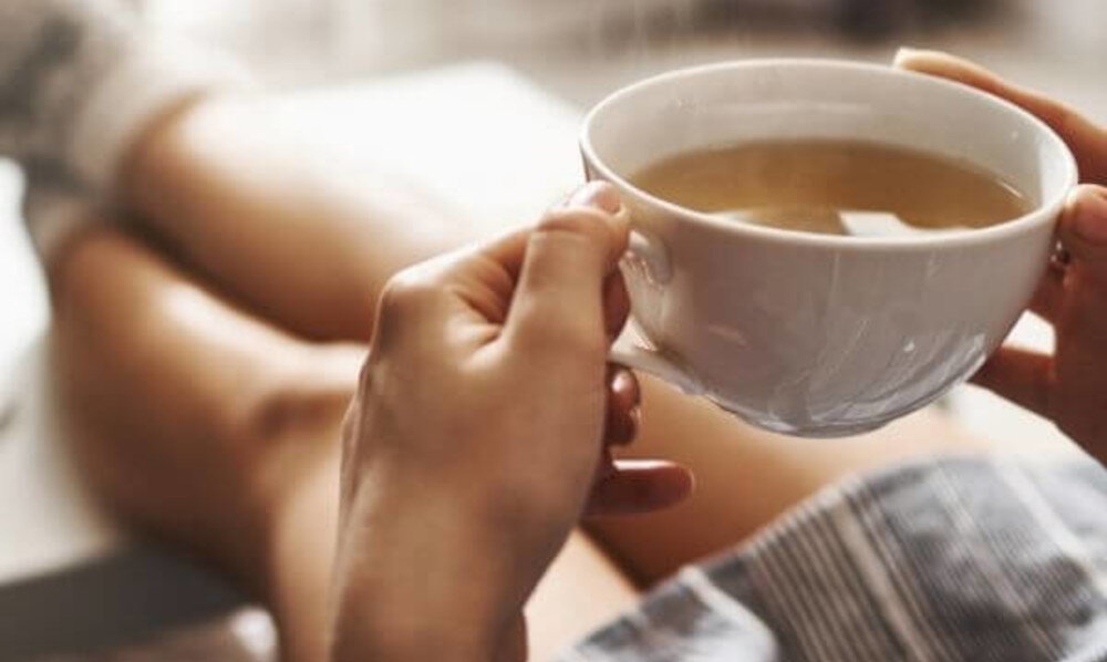 Osoba trzyma w dłoniach biały kubek z gorącym napojem. W tle widoczne są rozmyte nogi w pozycji siedzącej.