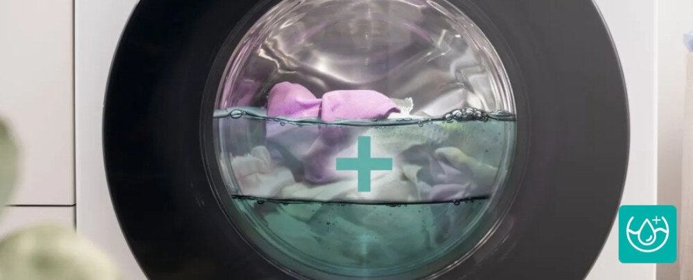 Widok na bęben pralki z wodą i kolorowymi tkaninami wewnątrz, na drzwiach widoczny jest symbol plusa. W prawym dolnym rogu znajduje się niebieska ikona z symbolem kropli i plusem.