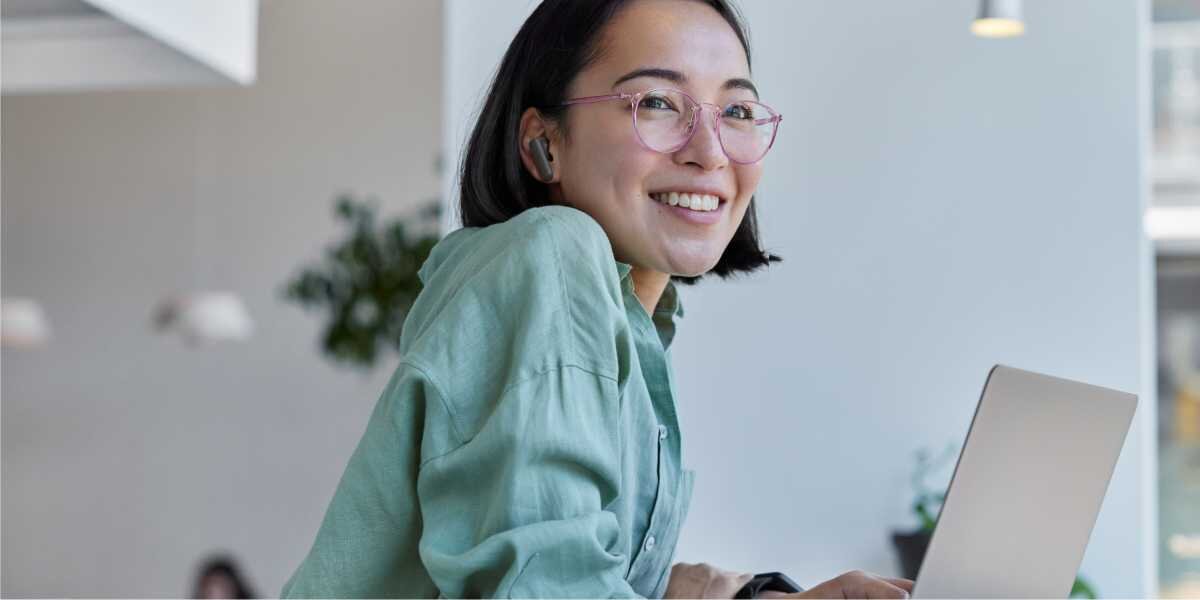 Słuchawki dokanałowe HAMA Spirit Touch ANC Czarny Uśmiechnięta kobieta w okularach i zielonej koszuli pracuje na laptopie w jasnym biurze, mając w uchu bezprzewodową słuchawkę douszną.